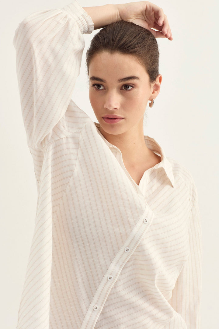 A woman wearing a cream-colored, long-sleeved button-up shirt with subtle vertical stripes, posed against a solid off-white background.
