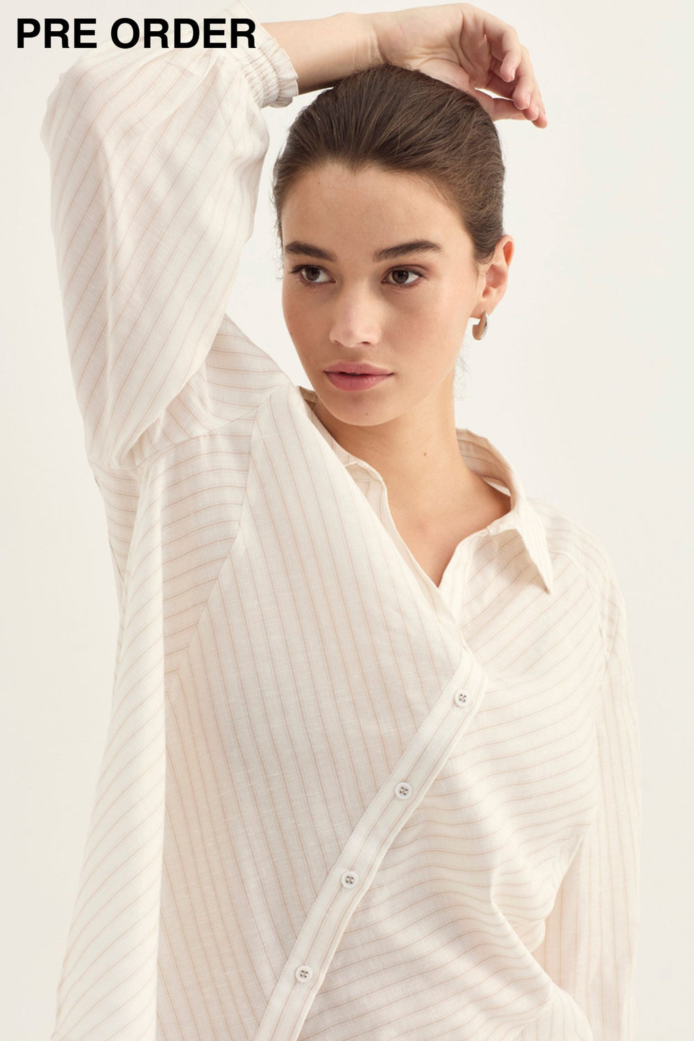 A woman wearing a cream-colored, long-sleeved button-up shirt with subtle vertical stripes, posed against a solid off-white background.