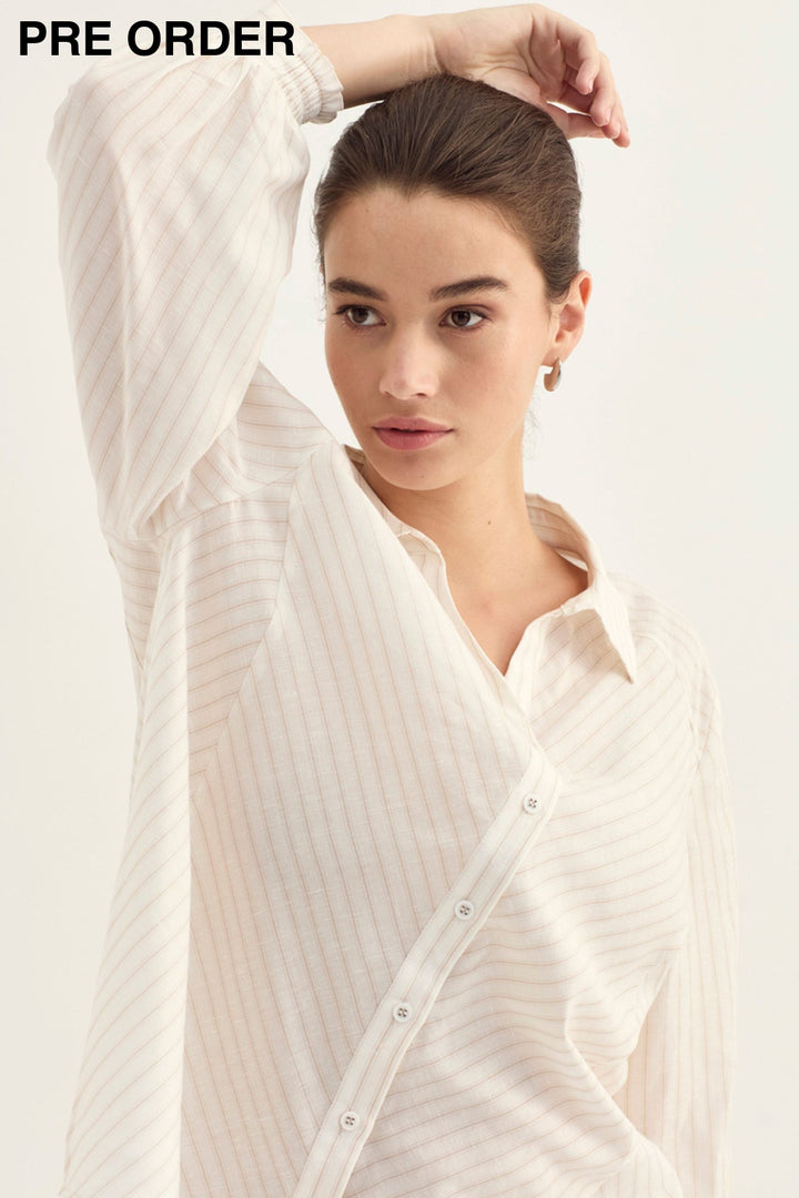 A woman wearing a cream-colored, long-sleeved button-up shirt with subtle vertical stripes, posed against a solid off-white background.
